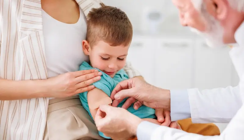 Γιατρός τοποθετεί επίδεσμο στο χέρι ενός μικρού παιδιού μετά από εμβολιασμό, με τη μητέρα του να το στηρίζει.