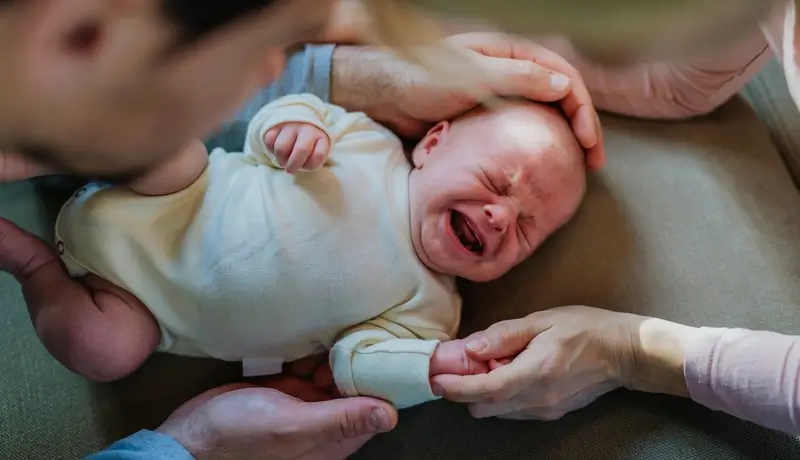 Βρέφος που κλαίει, ξαπλωμένο σε μαξιλάρι, ενώ δύο ενήλικες το παρηγορούν, κρατώντας το προσεκτικά.