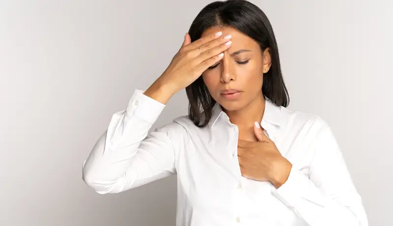 A woman in a white shirt holding her forehead and chest, appearing to experience discomfort or symptoms of illness.