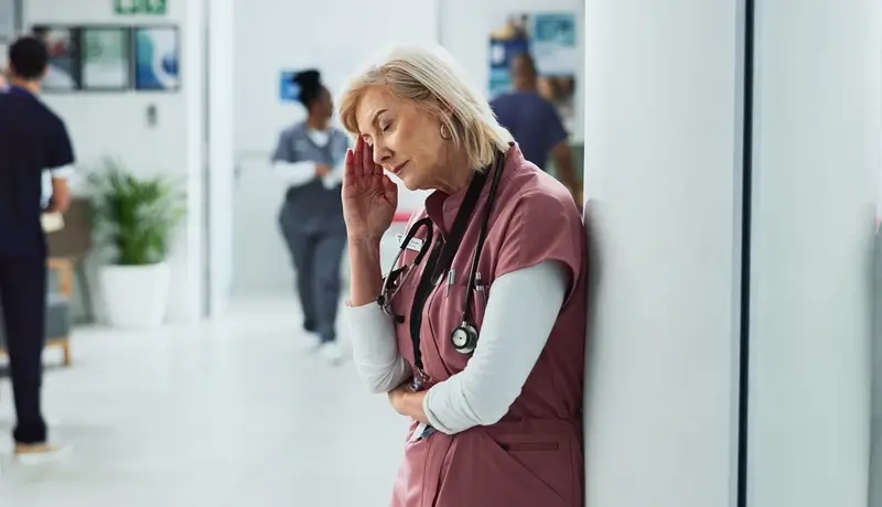 A tired female doctor in scrubs leaning against a wall, holding her head in fatigue in a busy hospital setting.