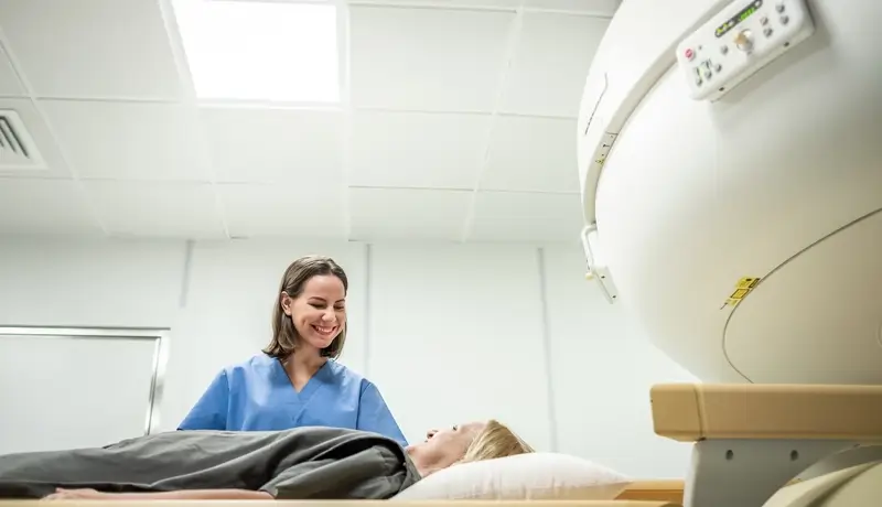 A patient lying on an MRI scanner table, smiling at a nurse in blue scrubs, inside a modern medical imaging facility.