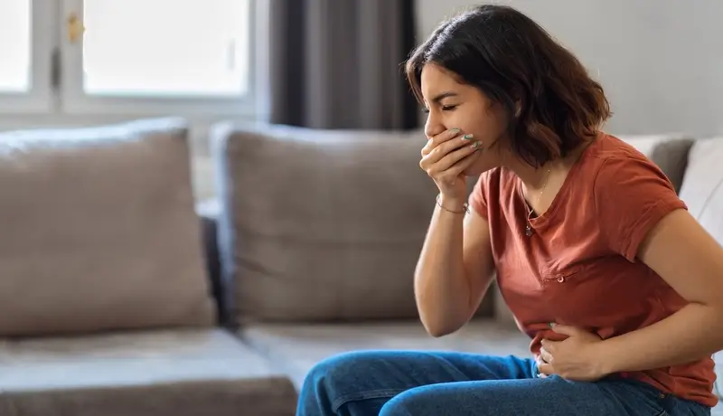 A woman sitting on a couch, holding her stomach and covering her mouth, showing signs of nausea and illness.