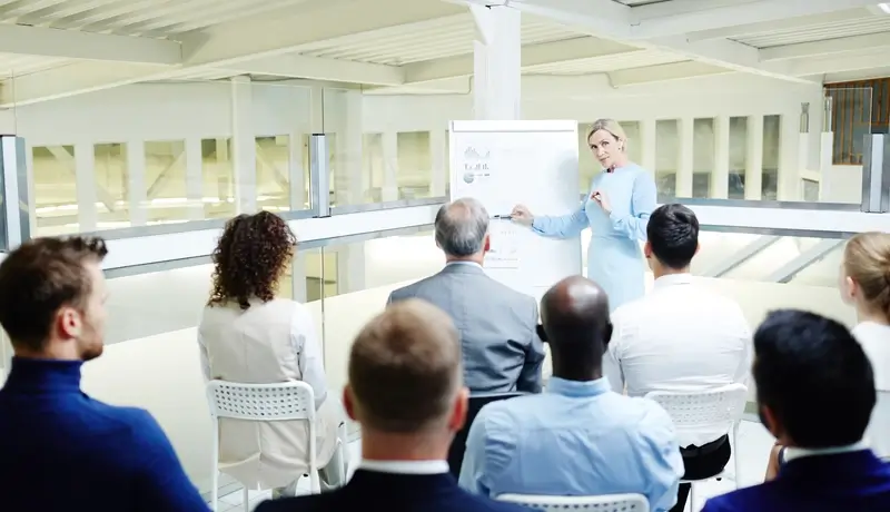 A professional woman presenting a training session to a diverse group of attendees in a modern conference room.