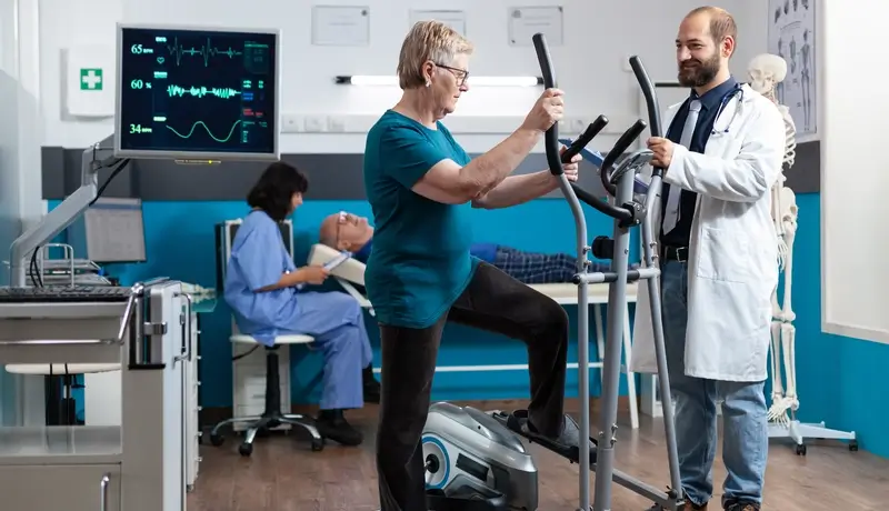 A senior woman using a stationary exercise machine under the guidance of a doctor, with medical staff and equipment visible in the background.