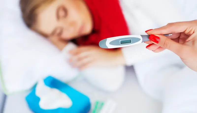 A person holding a thermometer in focus with a sick woman lying in bed blurred in the background.