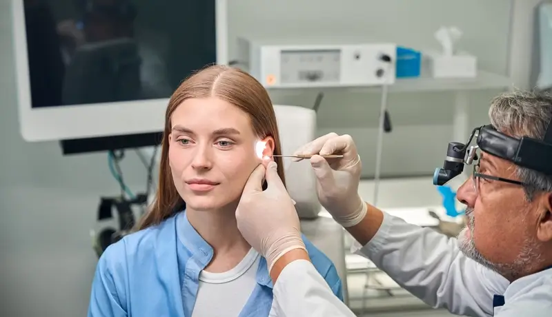 Μια γυναίκα υποβάλλεται σε εξέταση αυτιού από έναν γιατρό σε κλινικό περιβάλλον, χρησιμοποιώντας ειδικό φωτισμό και εργαλεία.