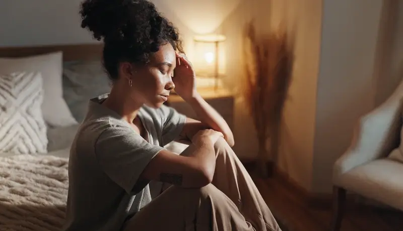 A woman sitting on the floor near a bed, leaning on her knees with a hand on her head, appearing thoughtful or distressed in a softly lit room.