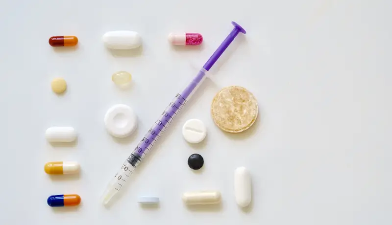 Μια σύριγγα τοποθετημένη ανάμεσα σε διάφορα φάρμακα και δισκία διαφορετικών σχημάτων και χρωμάτων, πάνω σε λευκό φόντο.