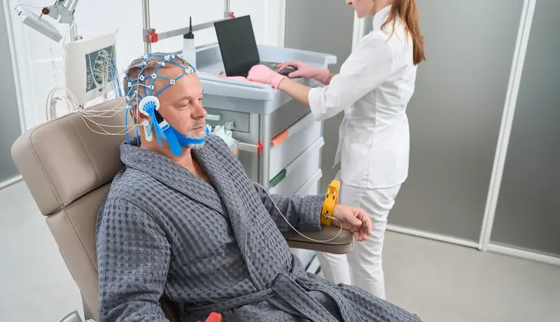 A man undergoing a medical procedure wearing a robe with electrodes attached to his head, while a medical professional operates nearby.