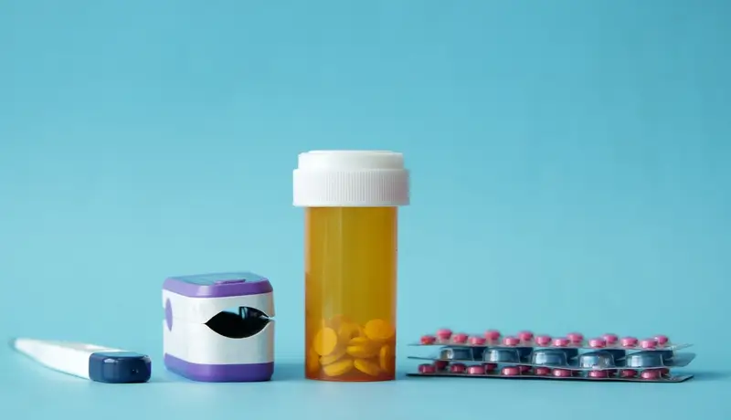 A collection of medical items, including a thermometer, pulse oximeter, pill bottle, and blister packs of medication, displayed against a light blue background.