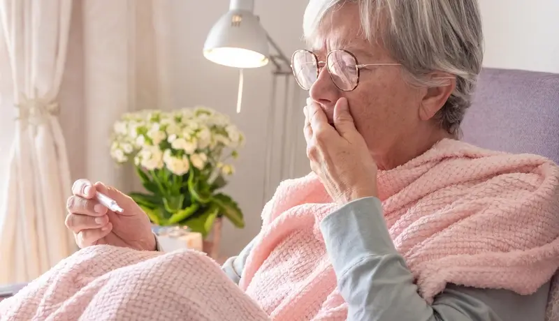 Ηλικιωμένη γυναίκα με συμπτώματα πνευμονίας, τυλιγμένη με κουβέρτα και καλύπτοντας το στόμα της καθώς βήχει.