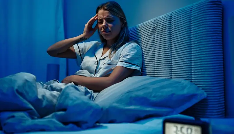A woman sitting in bed at night, holding her head with a pained expression, struggling with insomnia or discomfort, with an alarm clock displaying 3:50 AM.