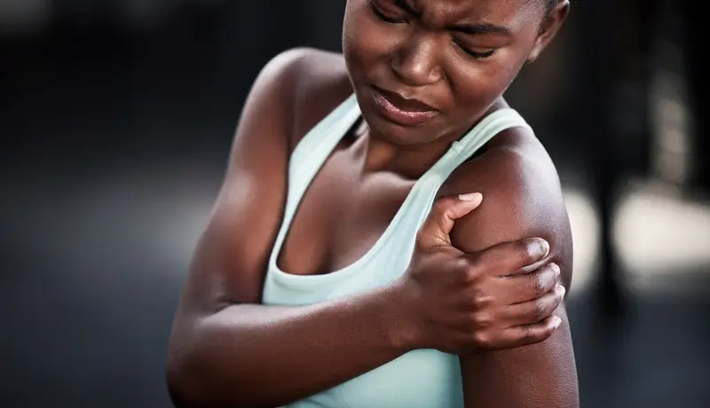 Γυναίκα που κρατά τον ώμο της, δείχνοντας σημάδια μυϊκού πόνου ή δυσφορίας στην περιοχή του ώμου.