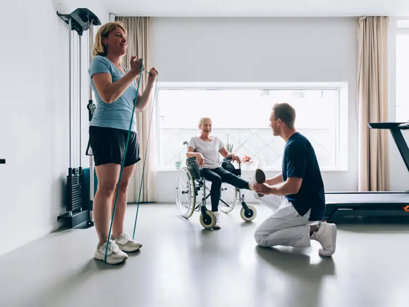 A rehabilitation specialist assisting a patient in a wheelchair while another patient performs resistance band exercises in a modern physical therapy clinic.