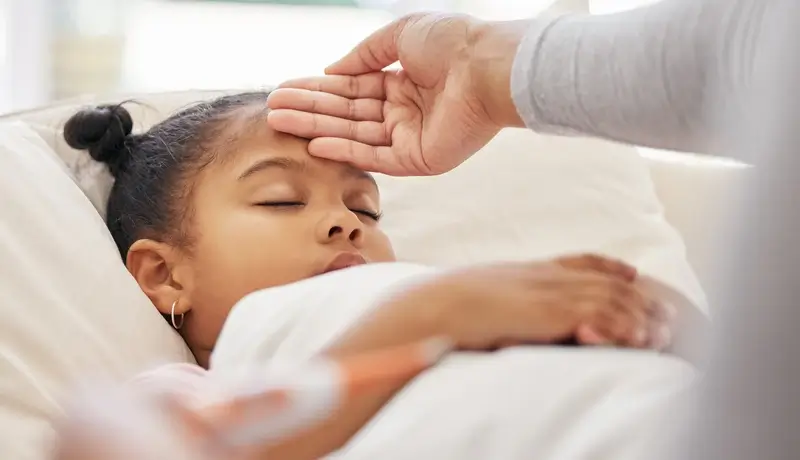 Παιδί που κοιμάται στο κρεβάτι, ενώ ένα χέρι ακουμπά στο μέτωπό του για να ελέγξει αν έχει πυρετό.
