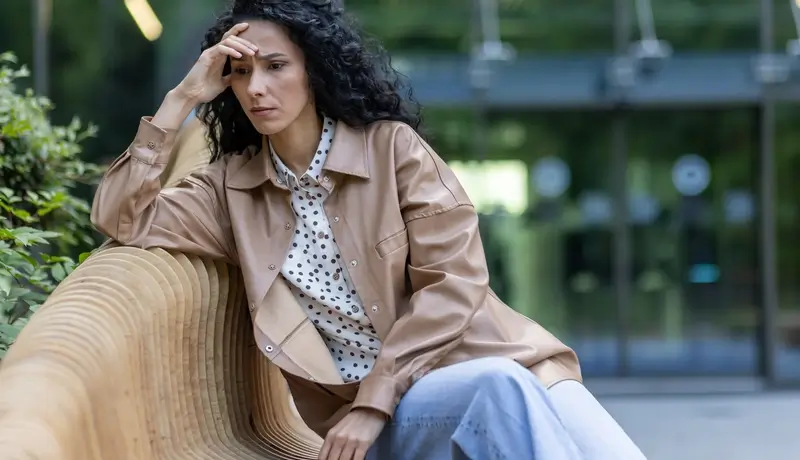 A woman sitting on a wooden bench outdoors, resting her head on her hand, appearing deep in thought or concerned.