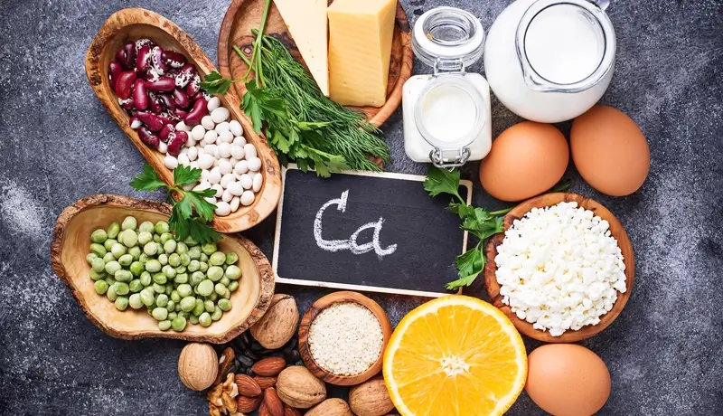 A variety of calcium-rich foods, including dairy products, eggs, legumes, nuts, seeds, and vegetables, displayed with a chalkboard labeled "Ca."