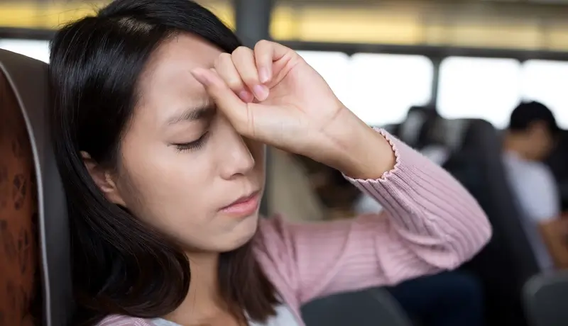 A woman sitting in a public transport seat holding her forehead, appearing to suffer from a headache and illness.